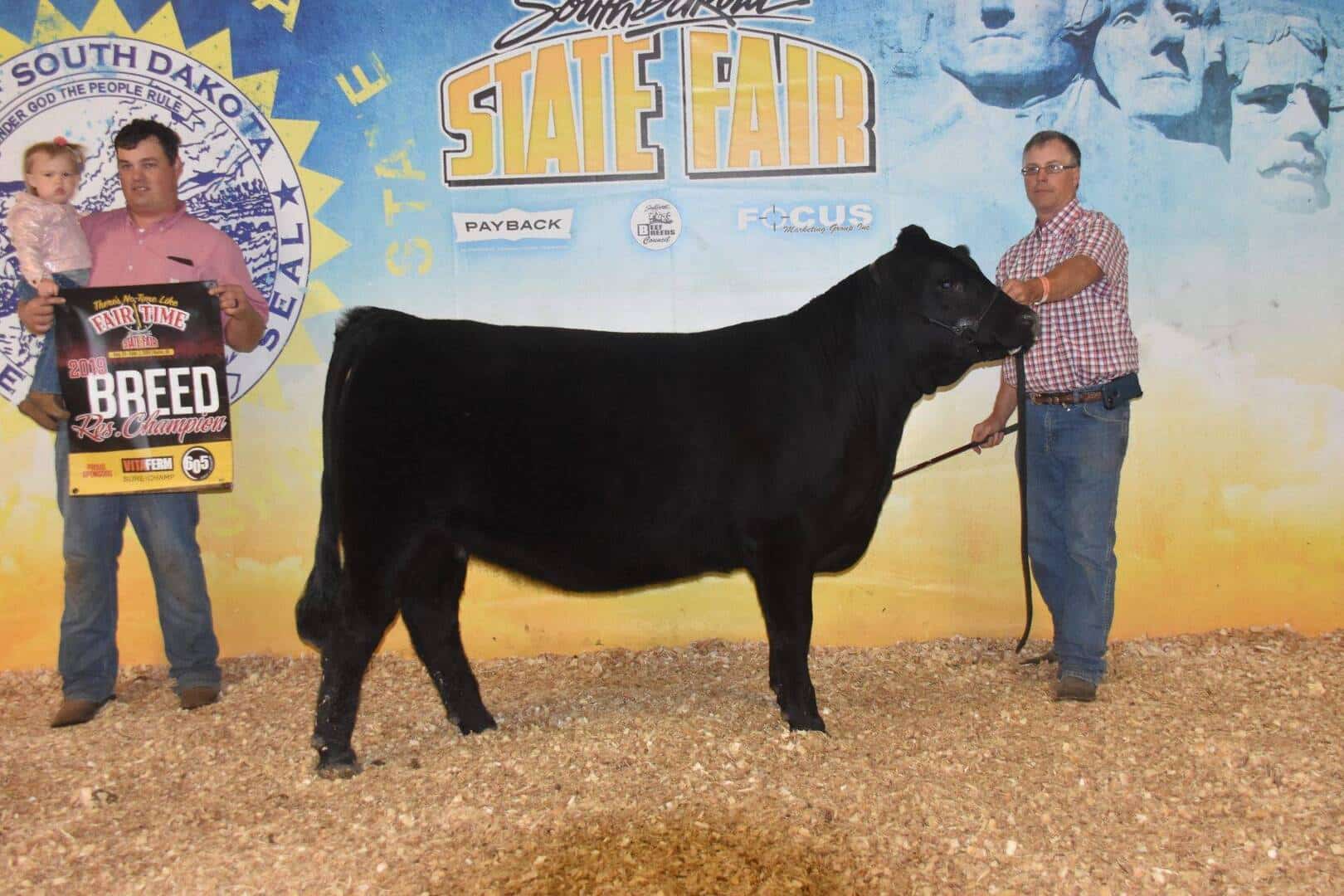 Two men stand on either side of a black cow at the South Dakota State Fair. One man holds a young child and a Breed Champion sign, while the other holds the cow’s halter. A fair banner is in the background.