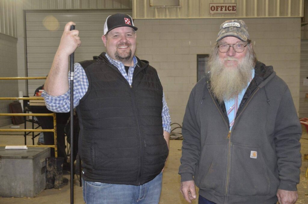 Two men stand indoors in a barn-like setting. One man wears a black vest, plaid shirt, and cap, smiling while holding a pole. The other, older man, has a long white beard, glasses, and a cap, wearing a jacket.