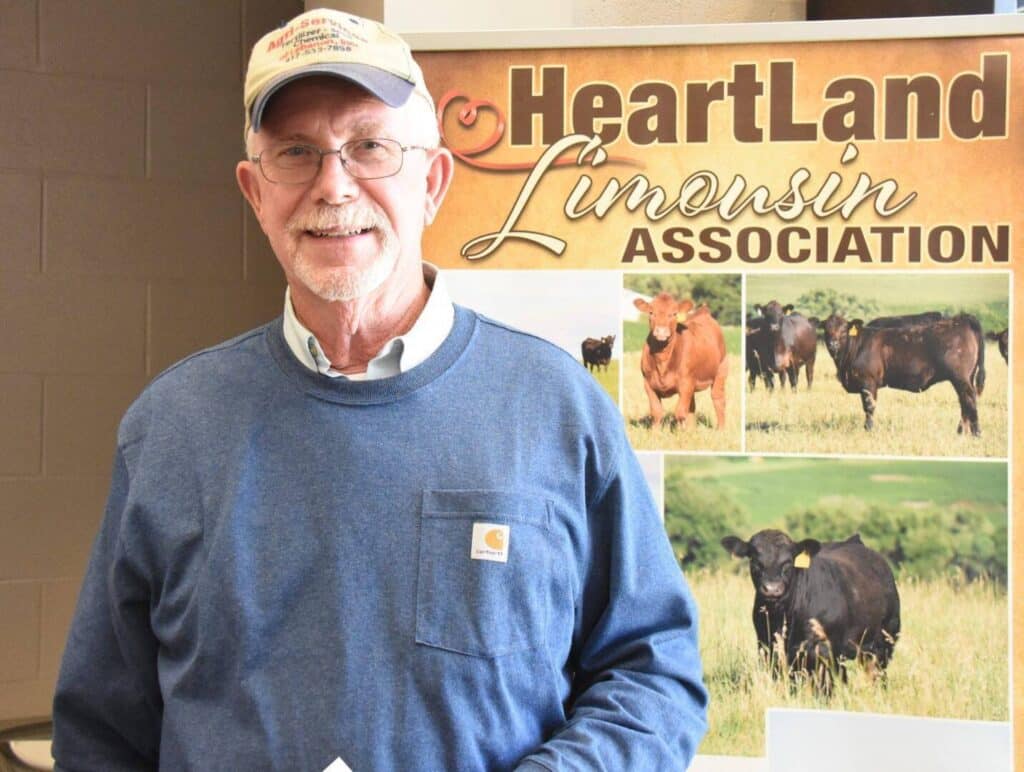 An older man wearing glasses, a beige cap, and a blue sweatshirt stands smiling in front of a Heartland Limousin Association sign with images of cows in a field.