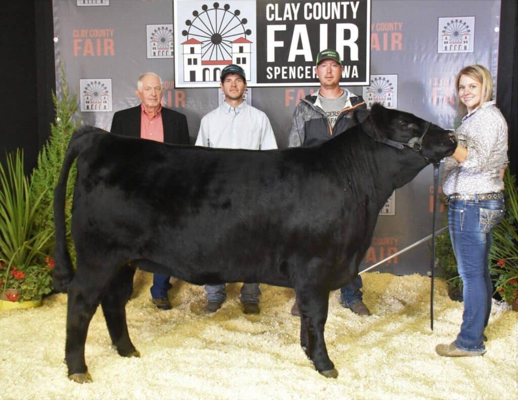 Four people stand behind a large black steer at the Clay County Fair in Spencer, Iowa, posing for a photo in front of a fair backdrop, with the handler holding the animal by a lead rope.
