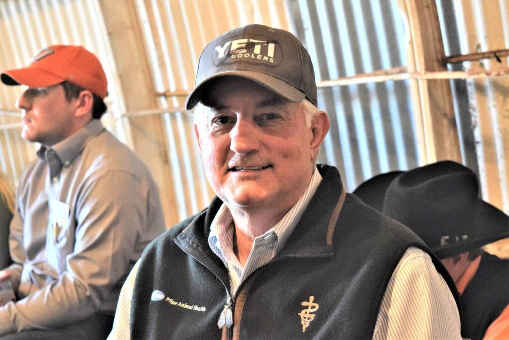 An older man wearing a YETI Coolers cap and a vest with a veterinary symbol smiles at the camera. Other people in casual clothing and cowboy hats sit in the background.