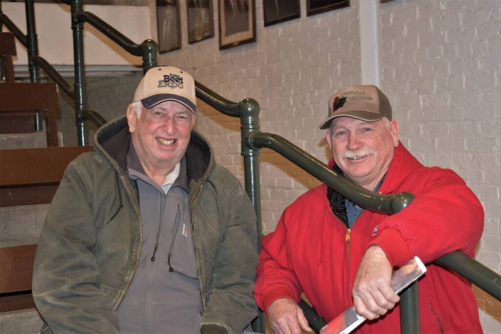 Two older men in jackets and baseball caps smile while sitting on stairs indoors. One wears a green jacket and gray cap, and the other a red jacket and tan cap. They appear cheerful and relaxed, with a brick wall behind them.