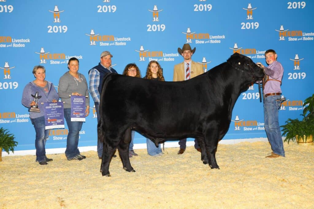 A group of seven people pose with a black steer and awards at the 2019 Houston Livestock Show and Rodeo, in front of a blue event backdrop.