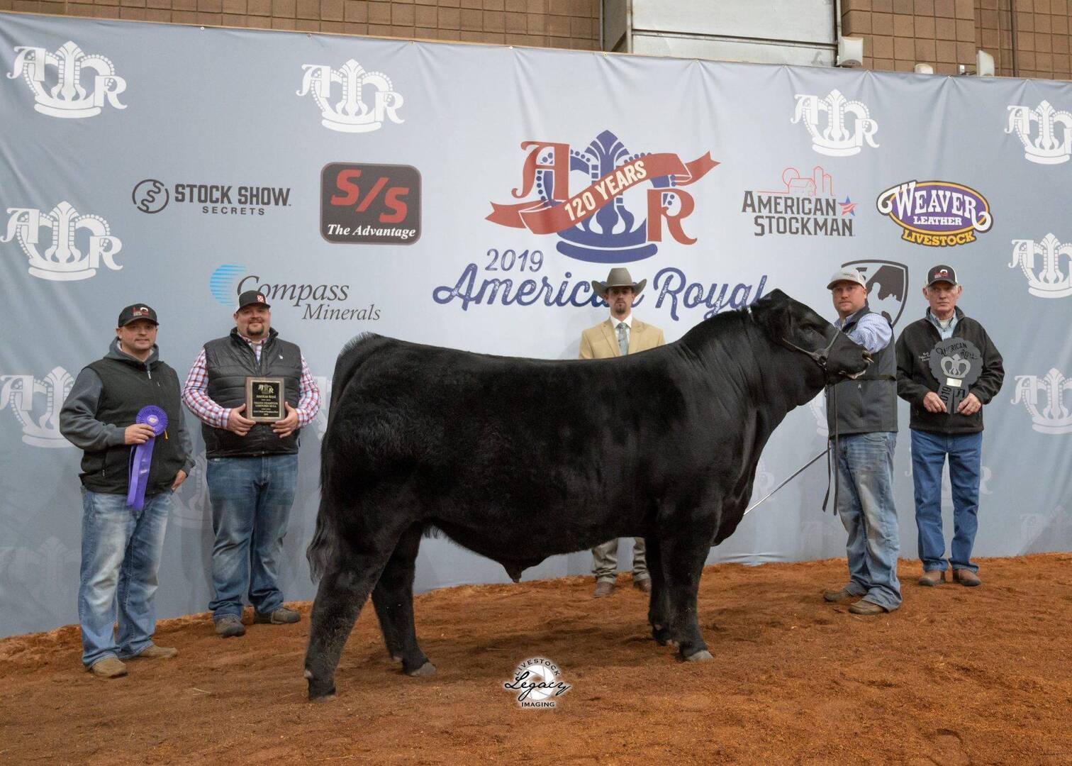Five people pose with a black cow at the 2019 American Royal livestock show. Three people hold awards and ribbons, and a branded backdrop with event logos is behind them on a red dirt floor.