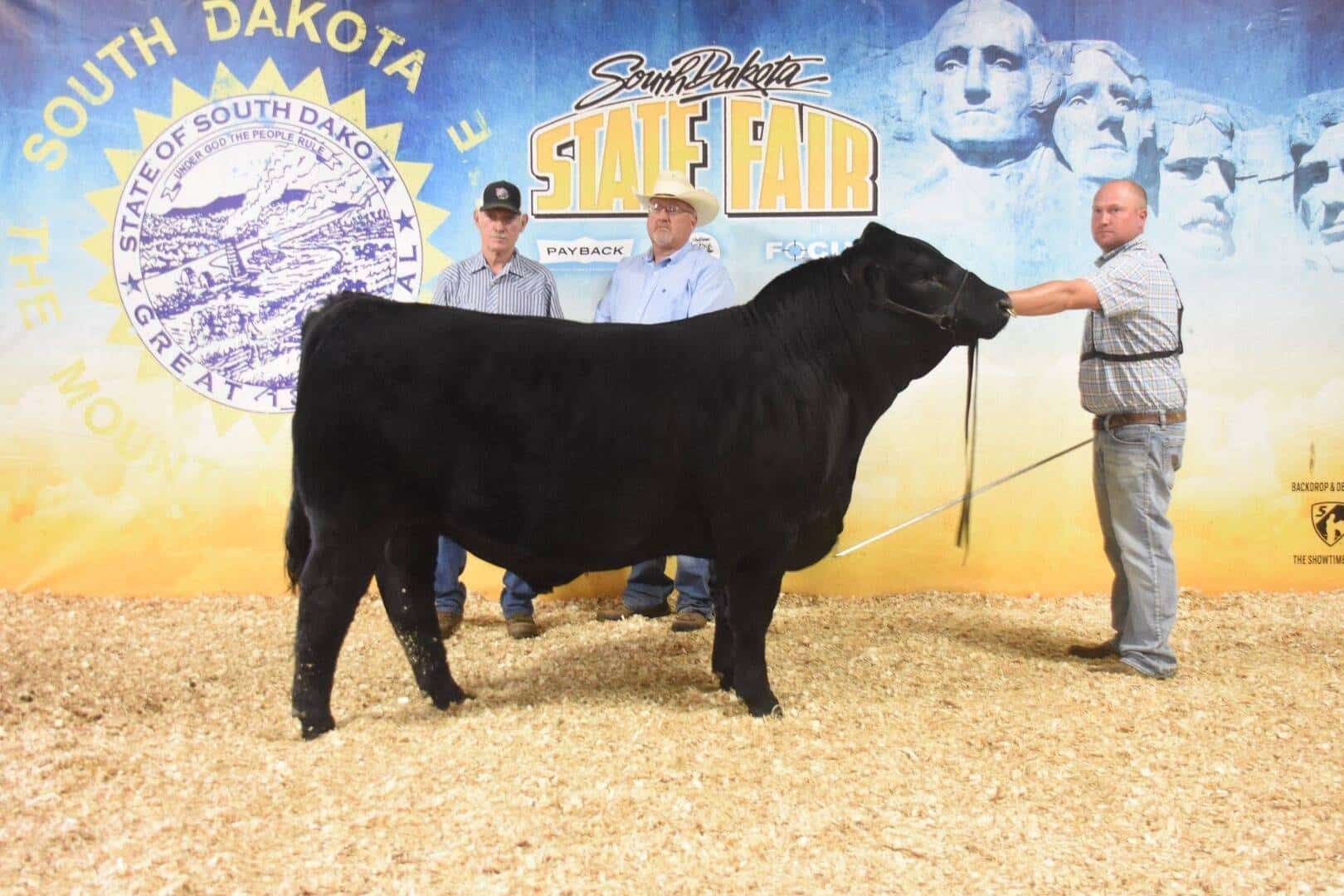 Three men stand with a black cow in front of a South Dakota State Fair backdrop featuring Mount Rushmore and the state seal. The men are wearing shirts, jeans, and hats, and the cow is being held on a lead.