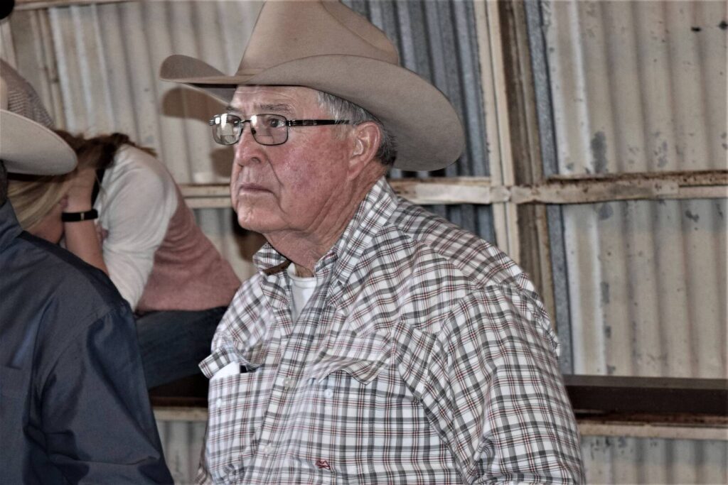 An older man wearing a cowboy hat, glasses, and a plaid shirt stands indoors in front of corrugated metal walls, looking to the side with a neutral expression.