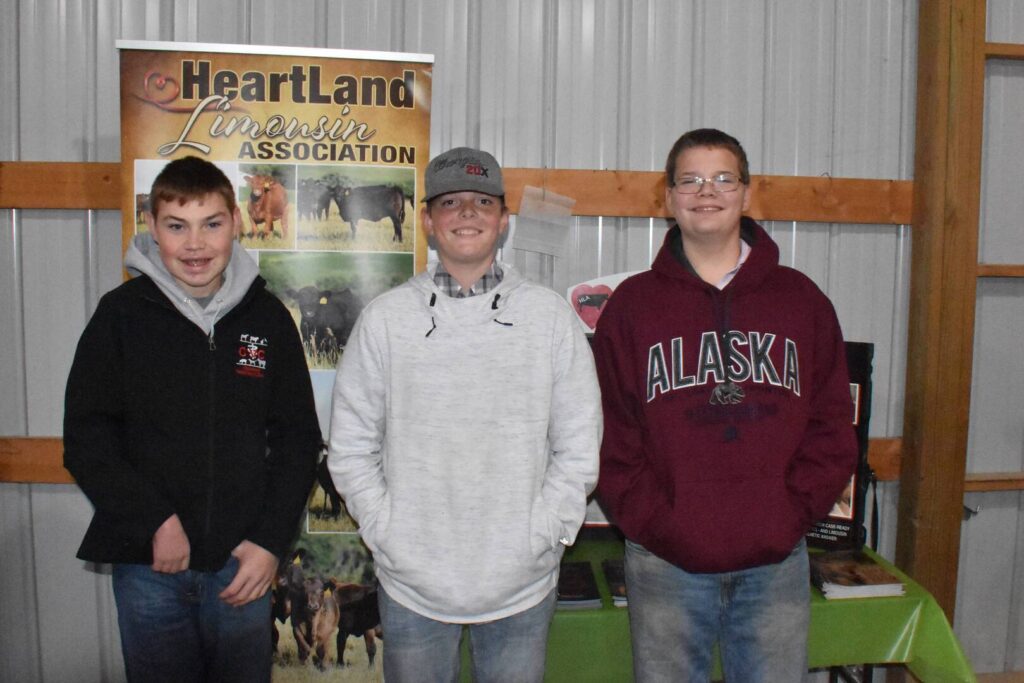 Three teenage boys stand indoors in front of a Heartland Limousin Association banner. They are smiling and wearing casual clothes, including hoodies and a hat. A table with pamphlets is visible behind them.