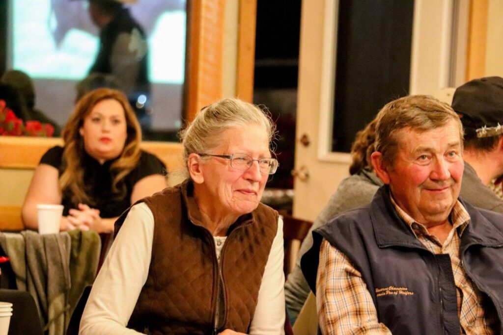 An older woman and man sit side by side, smiling, while other people sit in the background; the setting appears to be a casual indoor gathering or event.
