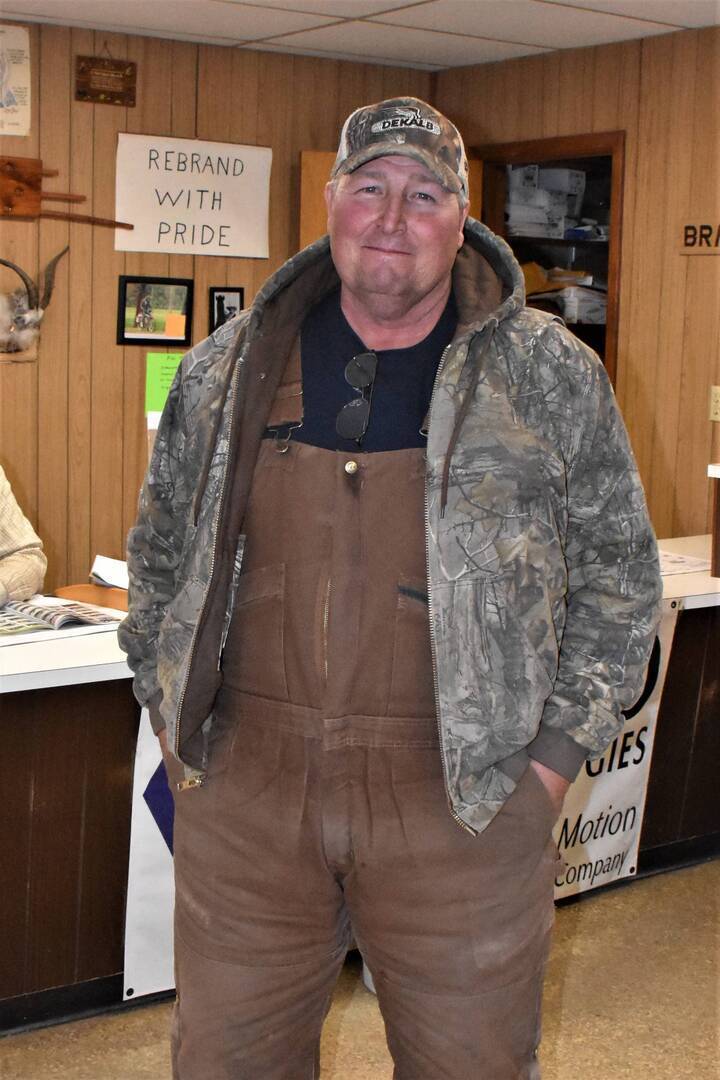 A man wearing a camouflage jacket, brown overalls, and a baseball cap stands indoors, smiling. Behind him, a wooden wall features a sign that reads REBRAND WITH PRIDE and various papers and photos.