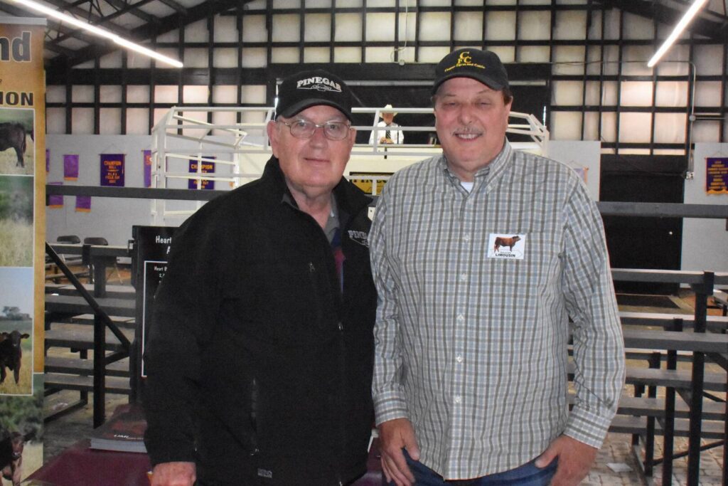 Two men wearing baseball caps and casual jackets stand side by side, smiling at the camera inside a barn or livestock exhibition area with pens and farm-related displays in the background.