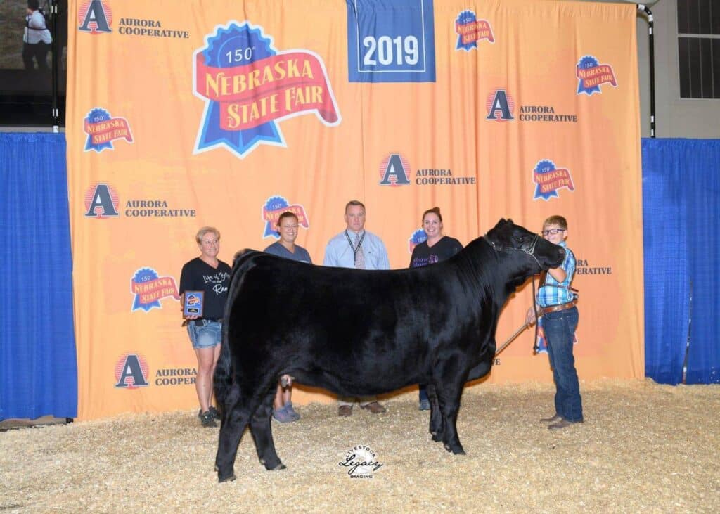 Five people pose with a large black cow in front of a Nebraska State Fair 2019 backdrop. The group stands on straw, smiling and holding awards, with blue curtains framing the scene.