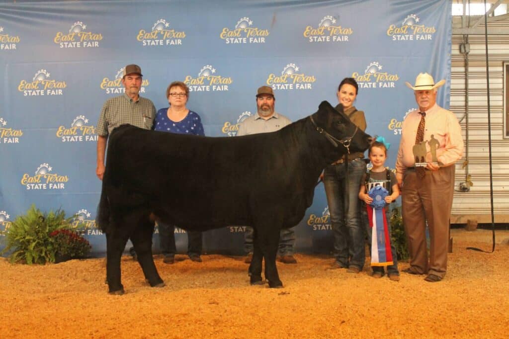 Six people stand behind a black cow at the East Texas State Fair. A young girl holds a trophy and ribbon, while an older man in a cowboy hat holds a plaque. They are on a dirt floor with a blue backdrop.