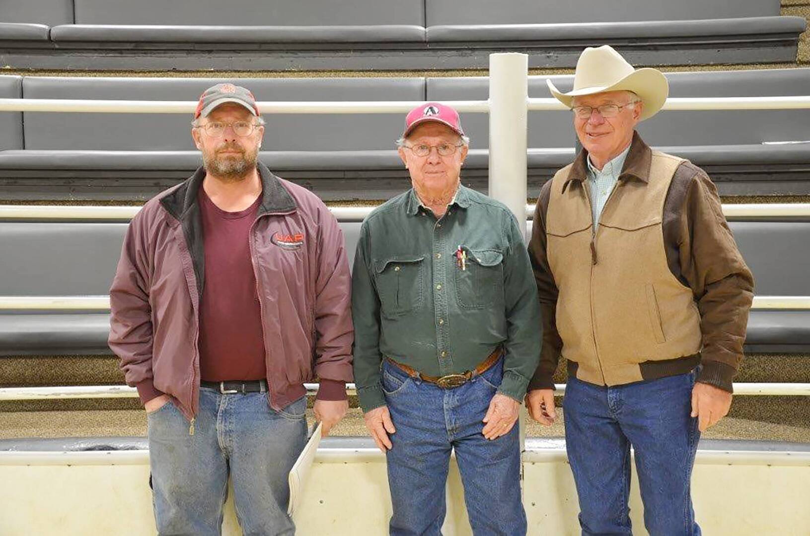 Three men stand side by side in front of tiered seating. They wear casual clothes: two in ball caps and glasses, and one in a cowboy hat and glasses. The setting appears to be an indoor arena or livestock venue.
