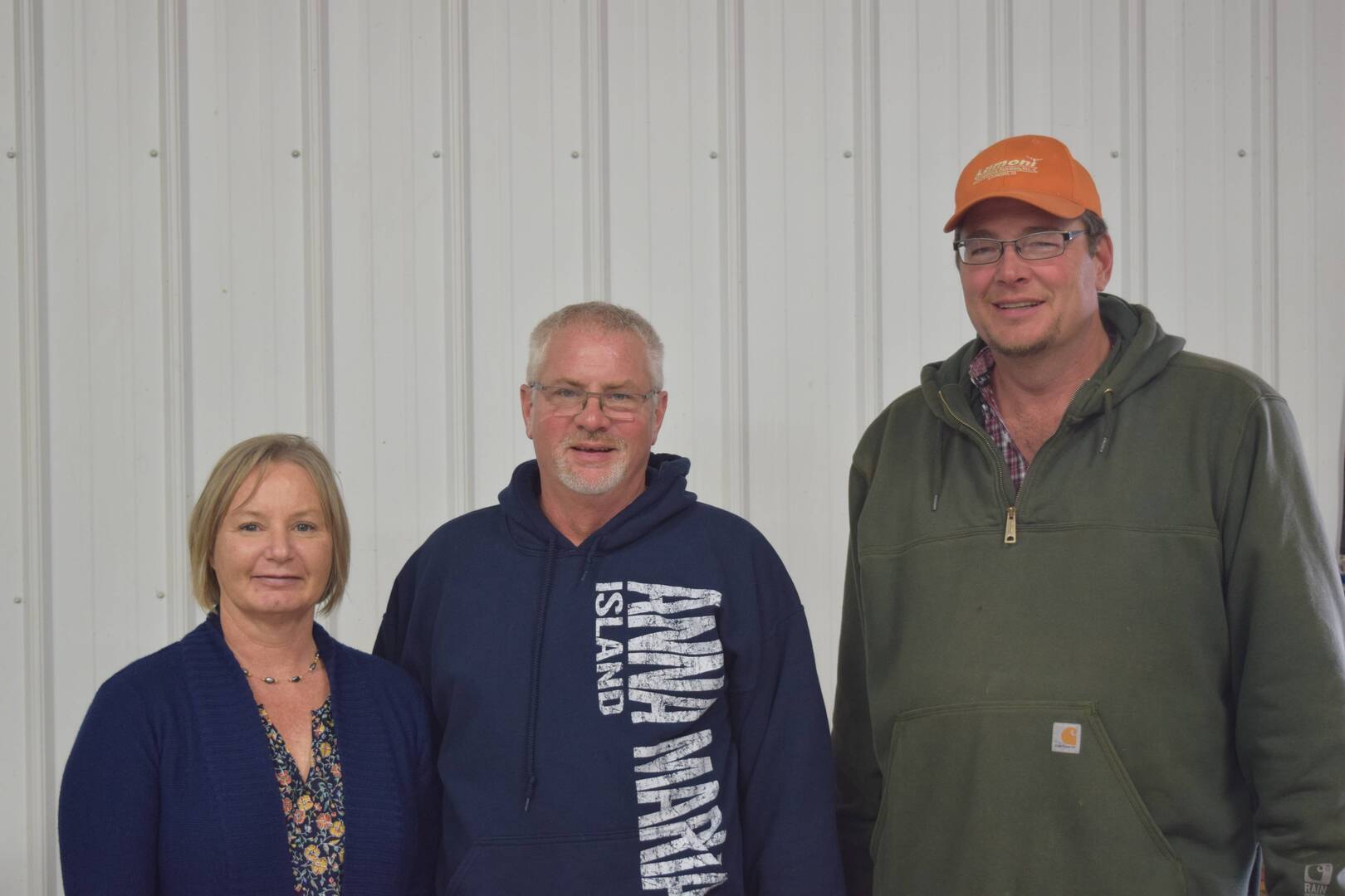 Three adults stand side by side in front of a white corrugated metal wall. The woman on the left wears a blue top, the man in the middle wears glasses and a blue hoodie, and the man on the right wears a green jacket and orange cap.
