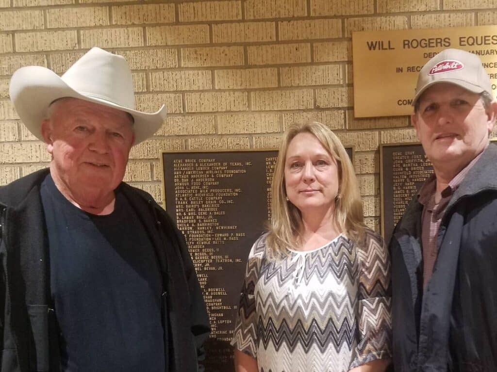 Three adults stand in front of a brick wall with plaques, including one reading Will Rogers Eques. The man on the left wears a cowboy hat, the woman in the center wears a patterned shirt, and the man on the right wears a cap and jacket.