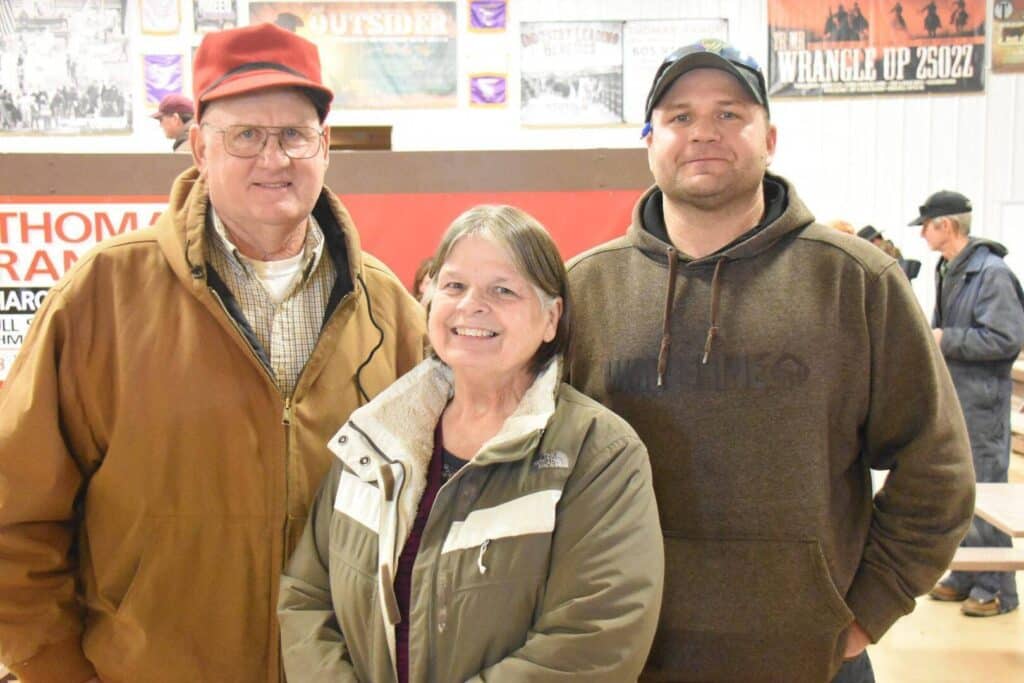 Three adults stand indoors, smiling at the camera. Two men wear brown jackets and hats, while a woman stands between them in a tan and green coat. Other people and posters are visible in the background.