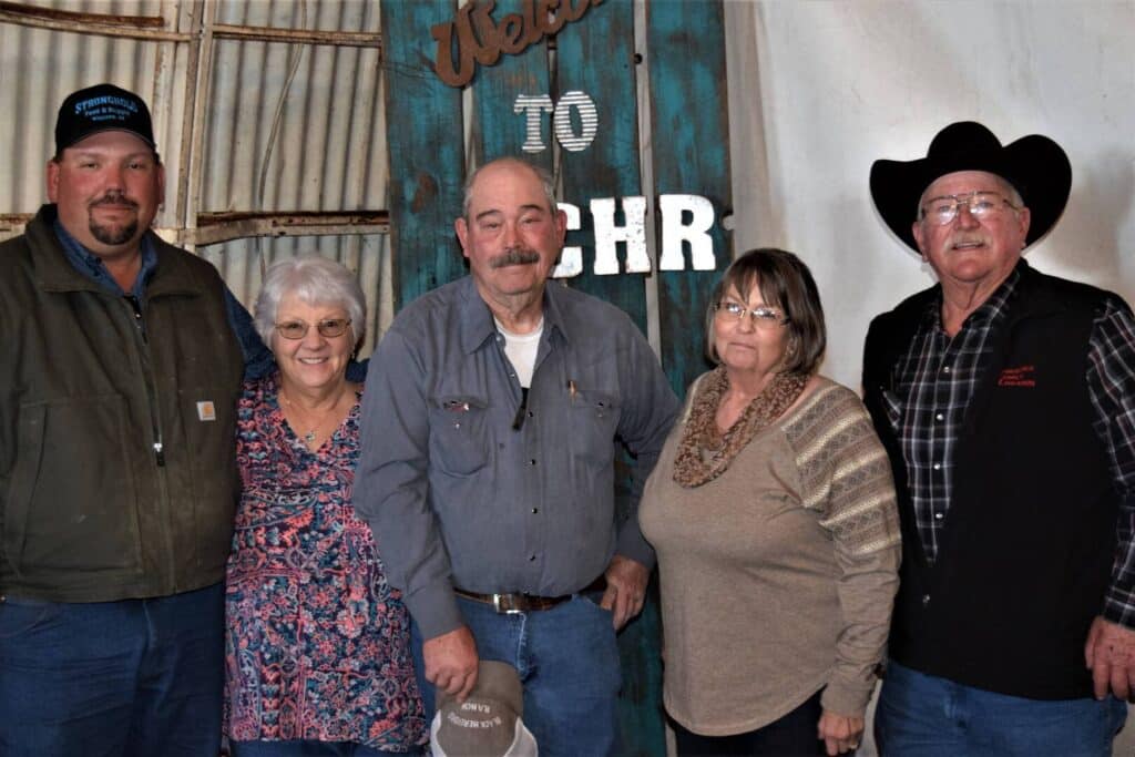 Five older adults stand side by side in front of a rustic Welcome to RCHR sign. Three men wear hats and casual clothes; two women stand between them, smiling. The group appears cheerful in a country-style setting.