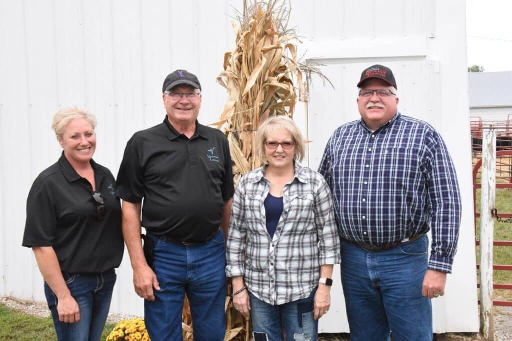 Four adults stand smiling in front of a white building and a tall bundle of dried corn stalks. They are wearing casual clothes, mostly jeans and collared shirts, with two men in baseball caps.