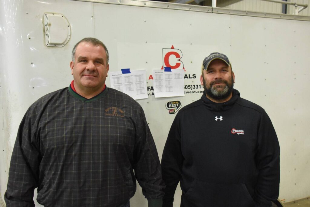 Two men stand side by side indoors in front of a white wall with printed papers and a red logo. Both are wearing dark sweatshirts; the man on the left has short hair, the man on the right has a beard and a baseball cap.