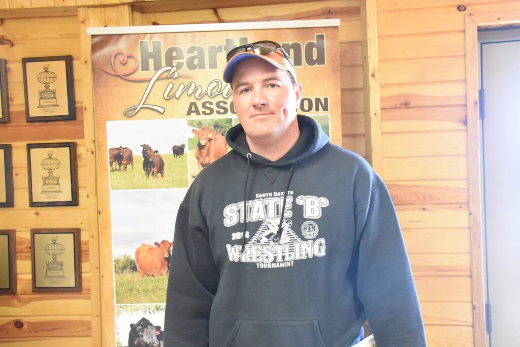 A man in a black hoodie and baseball cap stands indoors in front of a Heartland Limousin Association banner displaying cattle images, with wooden walls and framed awards in the background.