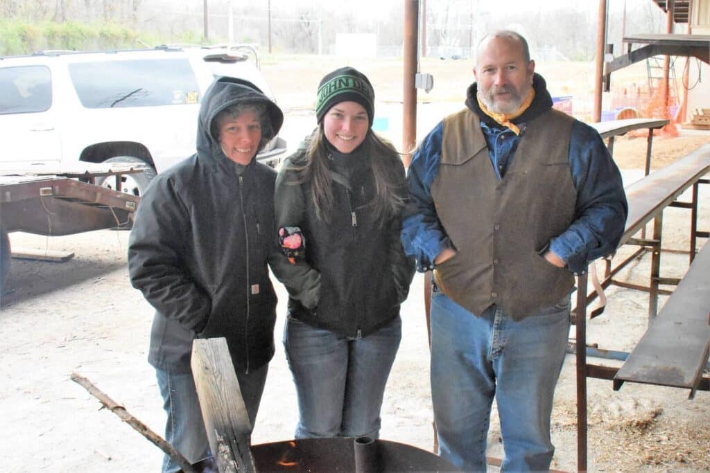 Three people dressed in warm clothing stand outdoors next to a fire pit, smiling at the camera. A white SUV and wooden picnic tables are visible in the background.