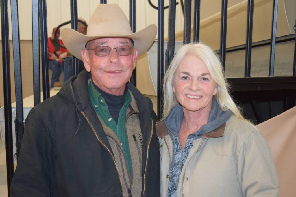 An older man in a cowboy hat and glasses stands next to an older woman with long white hair. Both are smiling and wearing jackets, posing indoors in front of black metal bars.