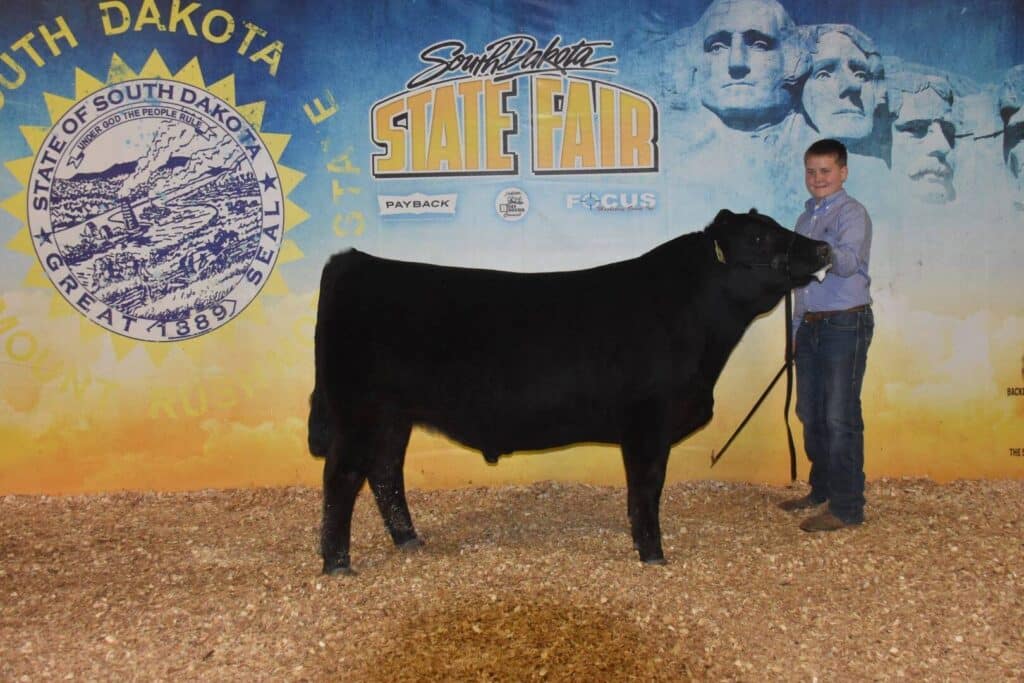 A person stands smiling next to a black cow at the South Dakota State Fair, in front of a backdrop featuring the state seal and Mount Rushmore.