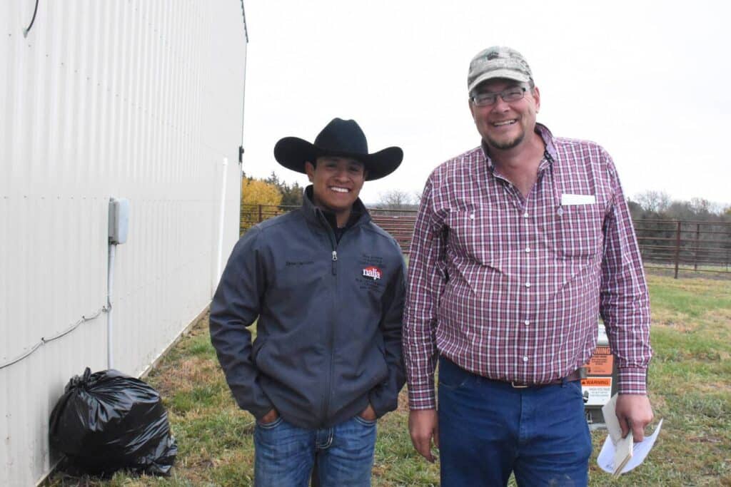 Two men stand outside by a metal building. One wears a black cowboy hat and gray jacket; the other wears glasses, a camo cap, and a plaid shirt. They are smiling, with grass and a wire fence in the background.