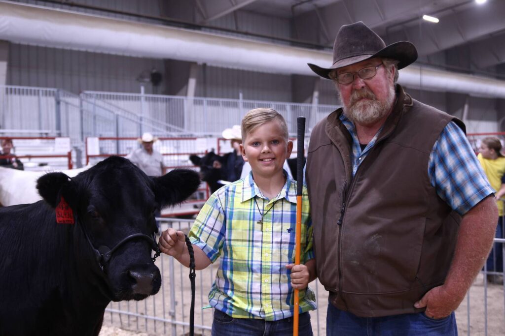 A boy and an older man stand next to a black cow inside a livestock show arena. The boy is holding a show stick, and both are smiling at the camera. Other people and cattle are visible in the background.