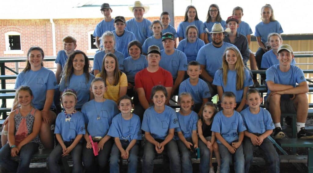 A large group of children, teens, and a few adults wearing matching blue T-shirts sit and stand on bleachers outdoors, smiling at the camera. One person in the center wears a red shirt.