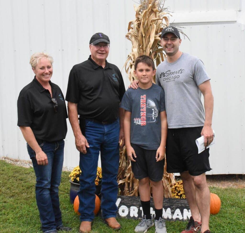 Four people stand in front of a white wall and a tall corn stalk decoration. Two adults are on each end, a child and another adult stand in the middle. They are smiling and standing on grass near pumpkins.