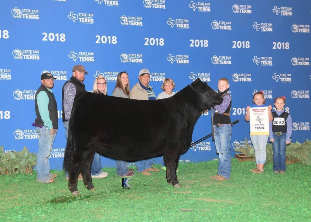 A group of people posing with a black cow at the State Fair of Texas 2018, standing in front of a blue backdrop with event logos; two children hold a Grand Champion sign and a ribbon.