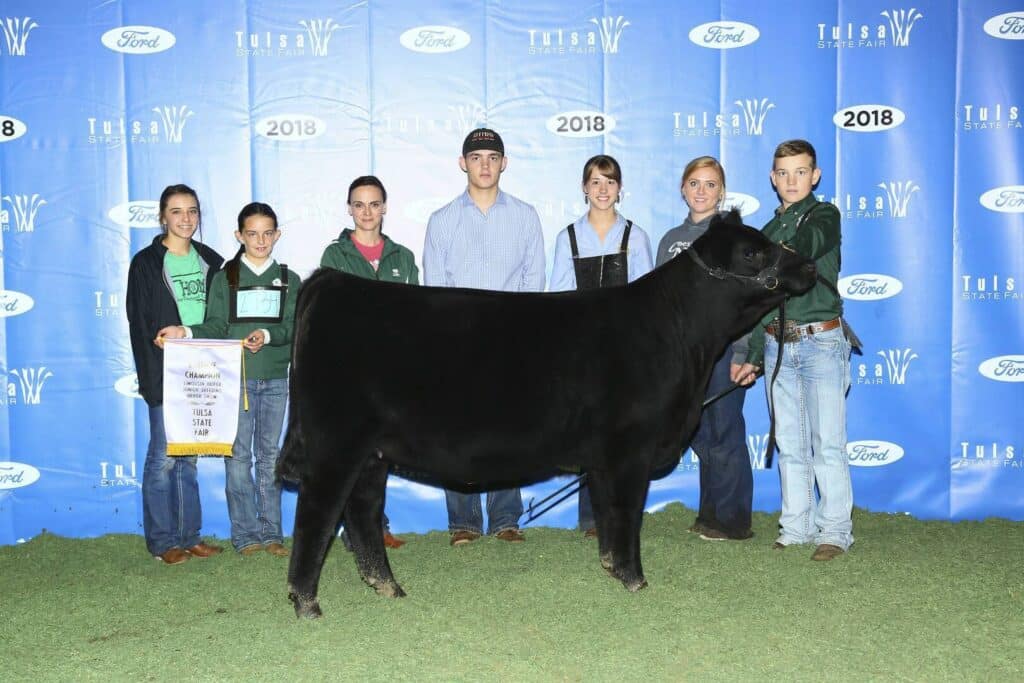 A group of six people pose with a black cow in front of a blue Tulsa State Fair 2018 backdrop. One person holds a banner and another holds the cows halter. The ground is covered with green turf.