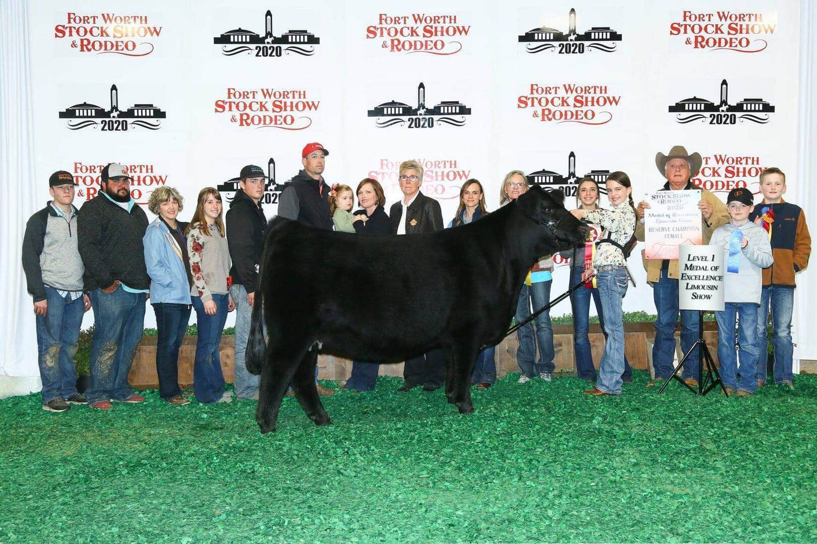 A group of people pose with a black cow indoors at the Fort Worth Stock Show & Rodeo 2020, standing in front of a step-and-repeat banner. One person holds an award sign next to the animal.