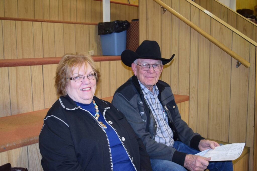 A smiling woman and an older man in a black cowboy hat sit side by side on wooden bleachers. The man holds some papers. They appear relaxed and are wearing casual jackets.