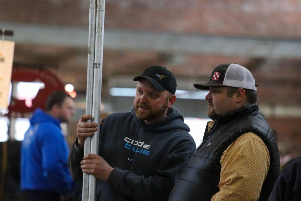 Two men in casual clothing and caps stand indoors, talking and looking in the same direction. One holds a vertical pole. The background is blurred, showing other people and a brick wall.