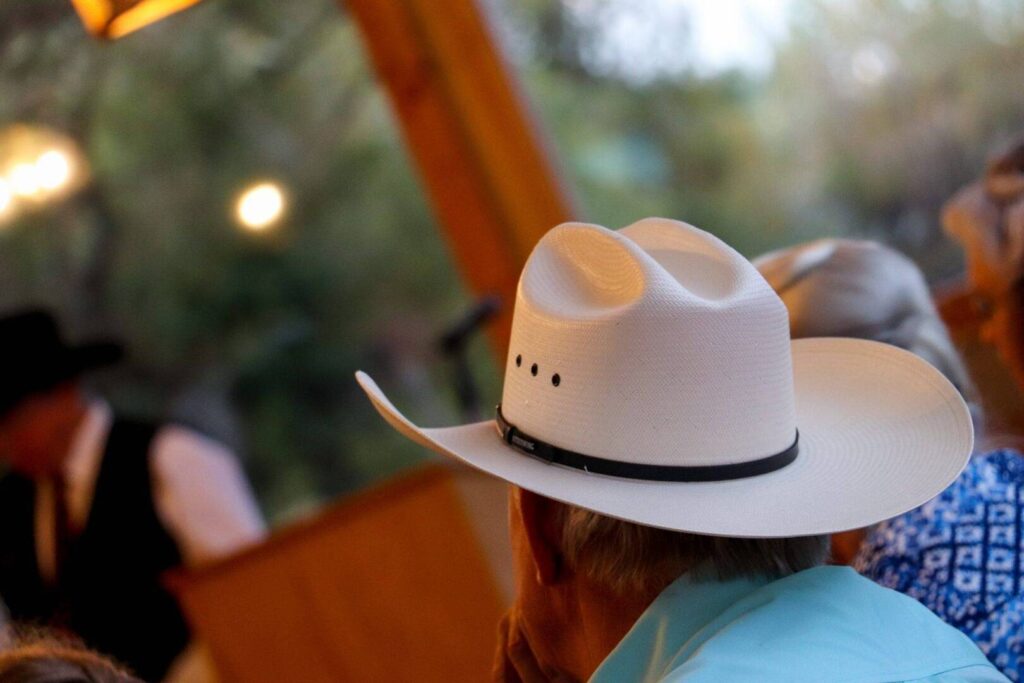 A person wearing a white cowboy hat sits facing away from the camera at an outdoor event, with blurred figures and trees in the background.