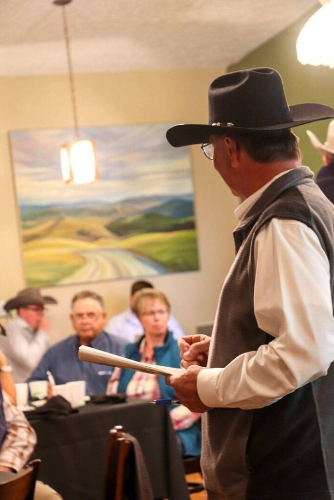 A man in a cowboy hat stands and speaks to a group of seated people in a room with a landscape painting on the wall. He holds a rolled-up paper, facing the group, who appear to listen attentively.