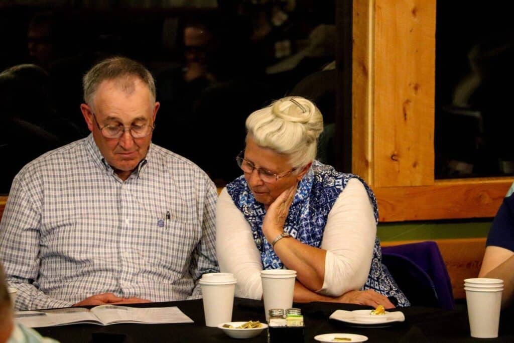 An older man and woman sit closely together at a table, reading a document. They both wear glasses and have gray hair. Several paper cups, plates with snacks, and condiments are on the table in front of them.