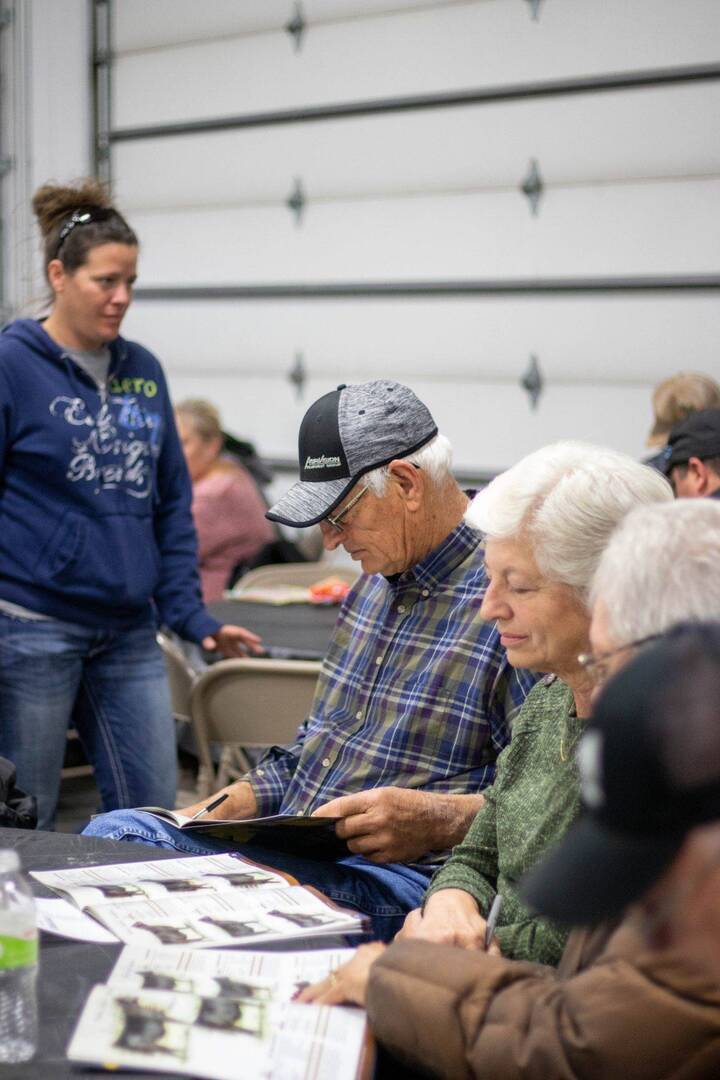 A group of older adults sit at a table in a casual indoor setting, looking at papers or booklets. In the background, a woman stands and others are seated. The atmosphere appears informal and social.