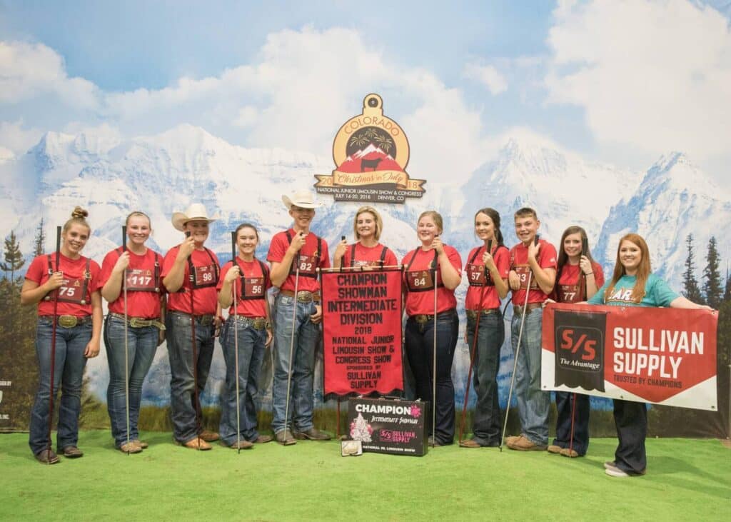 A group of young people in matching red shirts stand in front of a mountain backdrop, holding awards and banners for the Colorado State Fair, with Champion Showman Intermediate Division displayed.