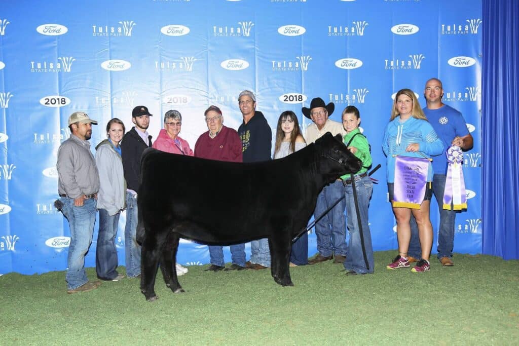 A group of people pose behind a black cow at a livestock show. A woman and boy hold a champion banner. The backdrop reads Tulsa State Fair 2018 with Ford logos. Everyone is smiling on green turf.
