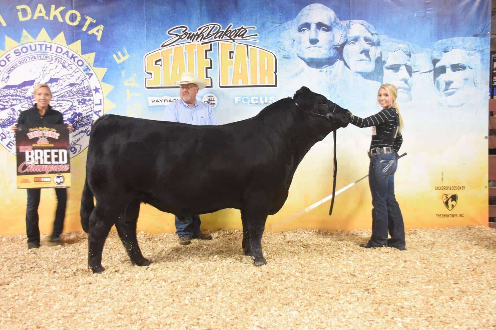 Three people pose with a large black bull at the South Dakota State Fair. The background features Mount Rushmore and event logos. One person holds a Breed Champion sign. The ground is covered in wood shavings.