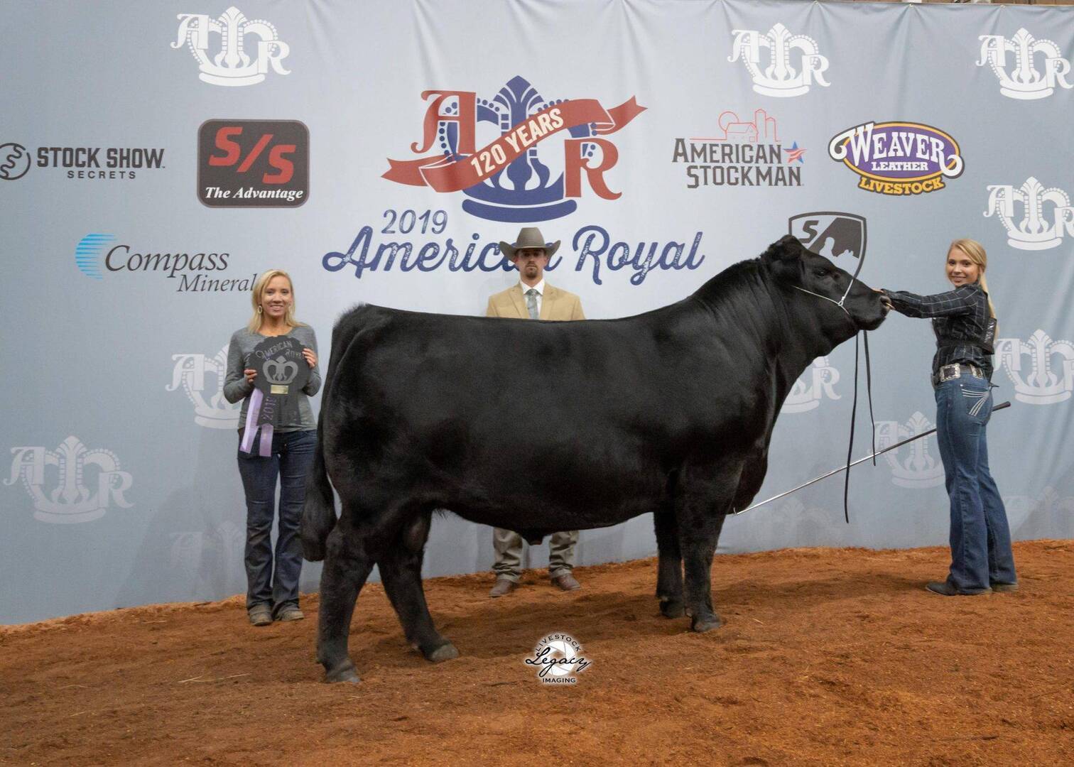 Three people pose with a black cow in front of an American Royal 2019 backdrop. The woman on the left holds a plaque, the man in the center wears a suit and hat, and the woman on the right holds the cow’s lead.