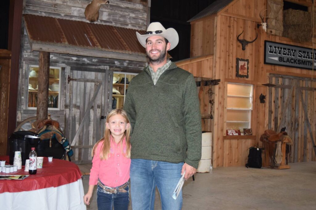 A man in a cowboy hat and a young girl stand smiling together indoors, in front of rustic wooden buildings with western decor. A table with drinks and a Livery Stable Blacksmith sign are visible in the background.