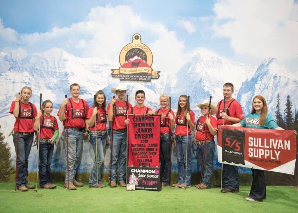 A group of young people in red shirts and jeans stand in front of a mountain backdrop, holding awards and a large banner reading Champion Showman Junior Division. An adult holds a Sullivan Supply sign on the right.
