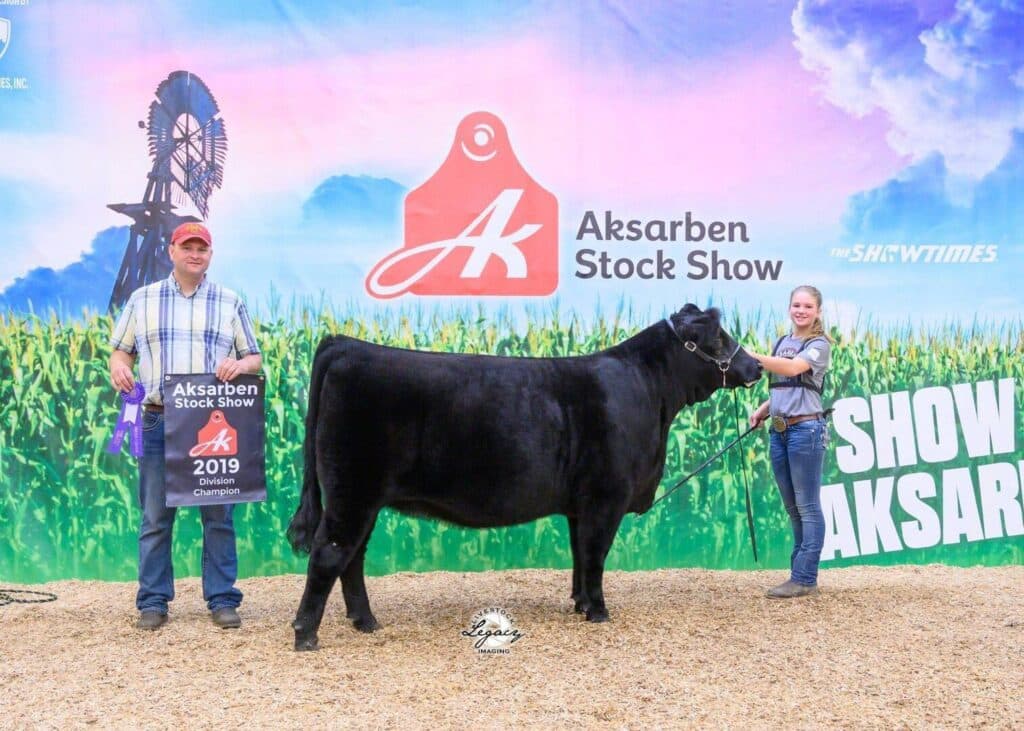 Two people pose with a black cow at the Aksarben Stock Show. One holds a banner reading 2019 Division Champion. A windmill and sponsor logos are visible on the colorful backdrop behind them.