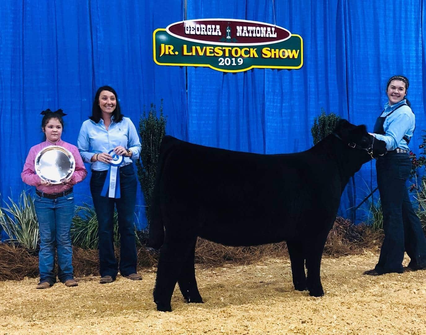 Three people stand next to a black cow in front of a blue curtain and a Georgia National Jr. Livestock Show 2019 sign. One holds a ribbon, another a silver platter, and the third holds the cow’s halter.