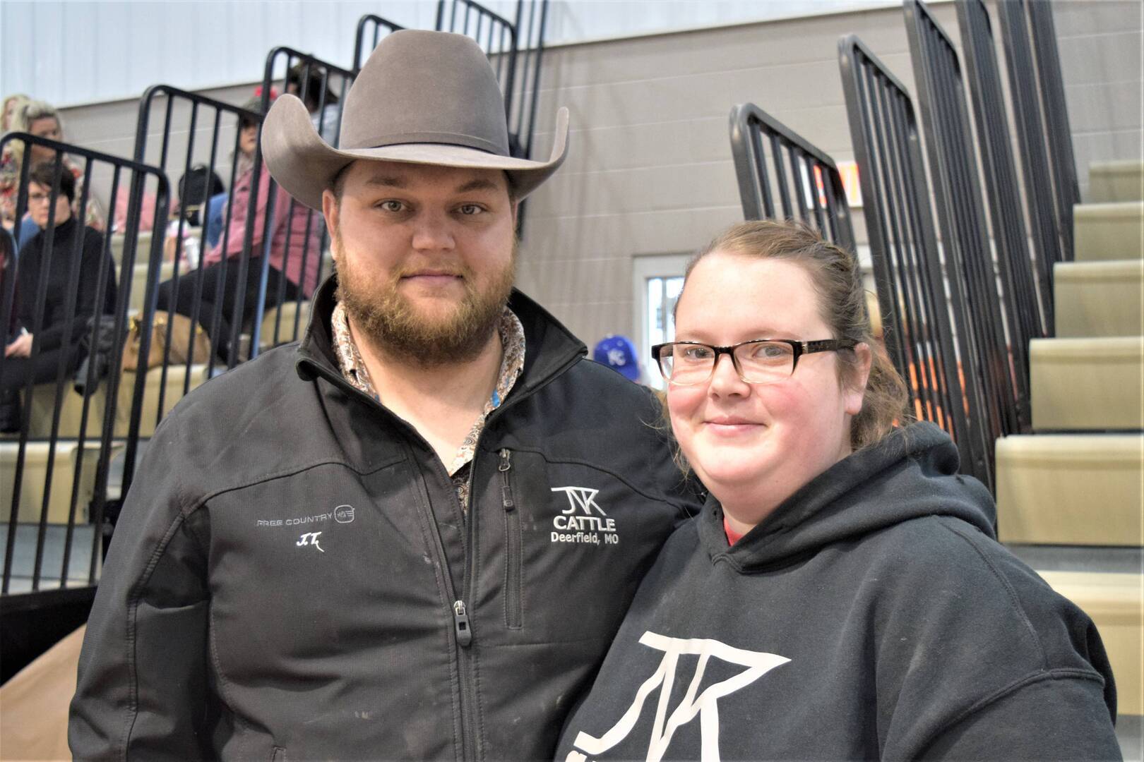 A man wearing a cowboy hat and a black jacket stands next to a woman in glasses and a black hoodie, both smiling, with bleachers and people in the background.