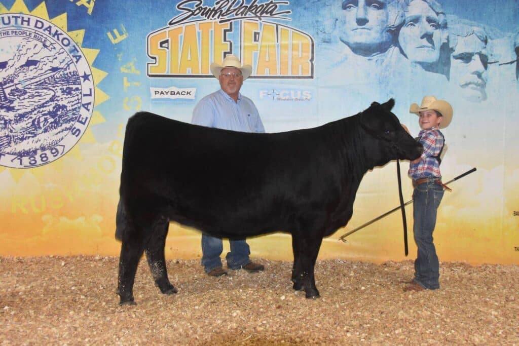 Two people in cowboy hats stand beside a black cow at the South Dakota State Fair. They pose for a photo in front of a fair-themed backdrop with Mount Rushmore and fair logos visible.